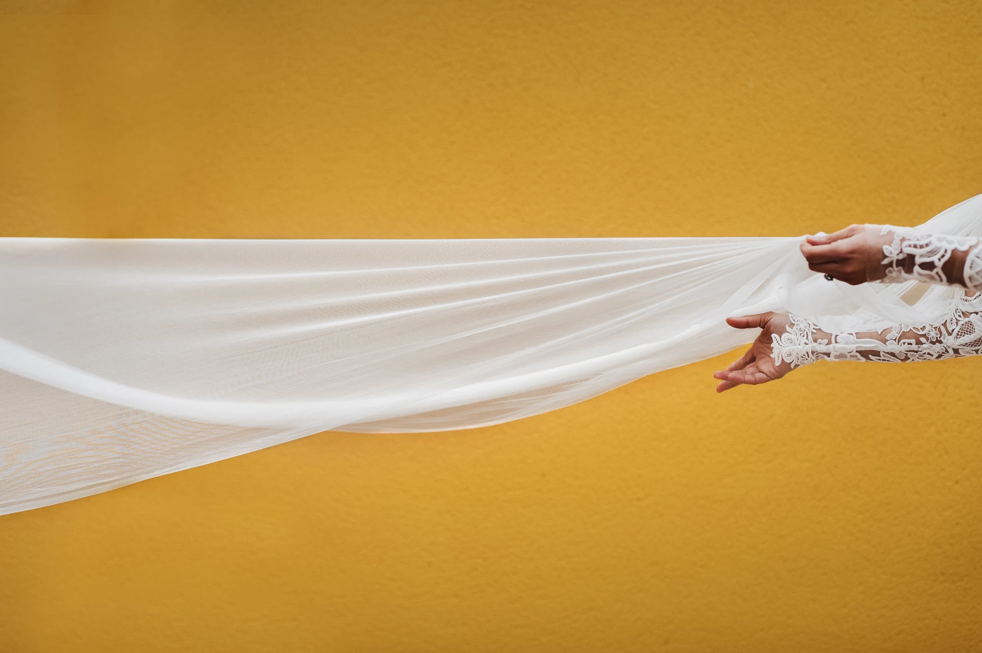 Bride's lace sleeve holding a long tulle veil streaming across a golden yellow wall
