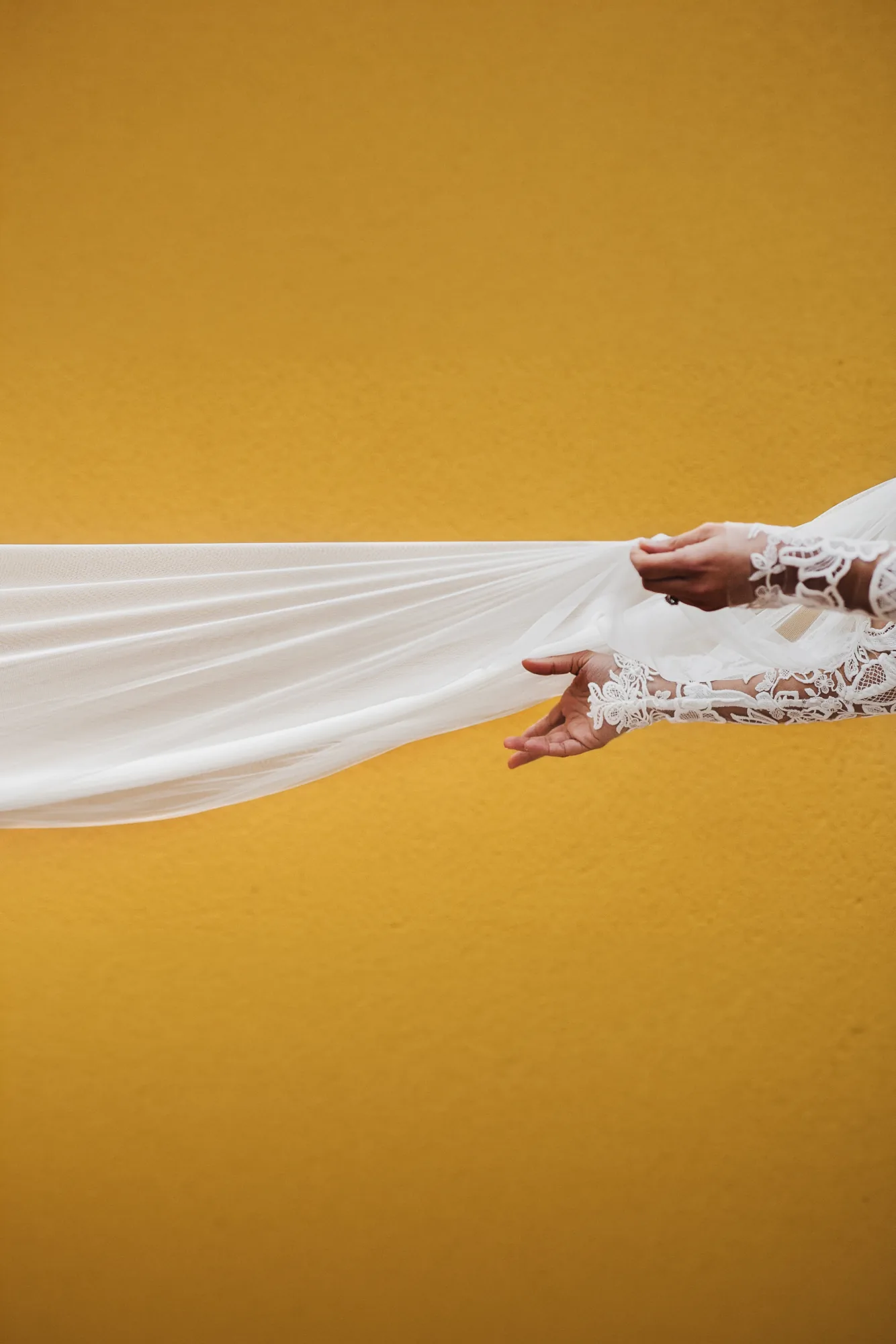Bride's lace sleeve holding a tulle veil across a golden yellow wall