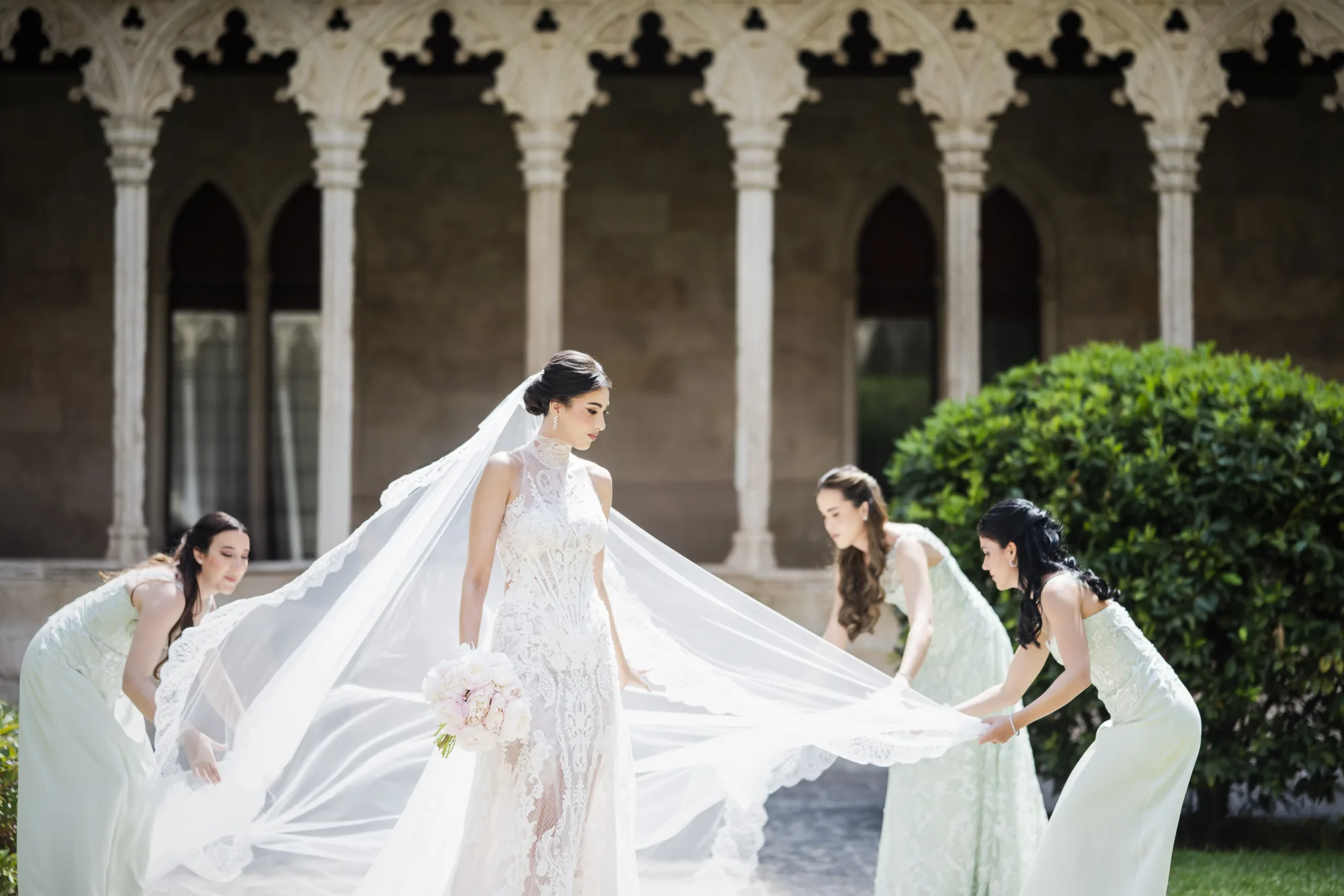 Bridesmaids spreading the bride's long veil in front of an arched cloister