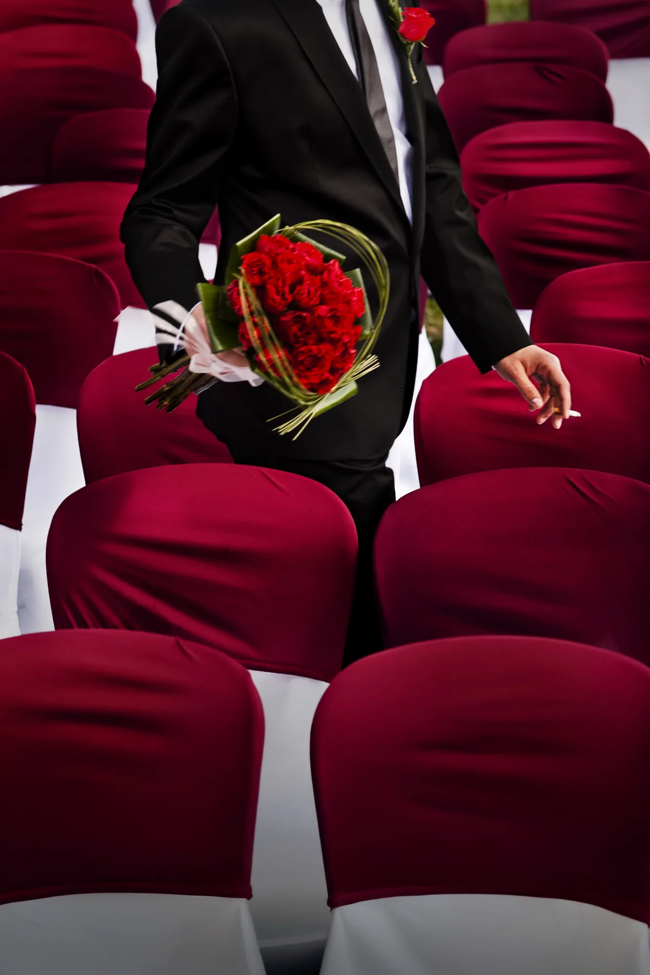 Groom holding a red rose bouquet behind his back among red seats